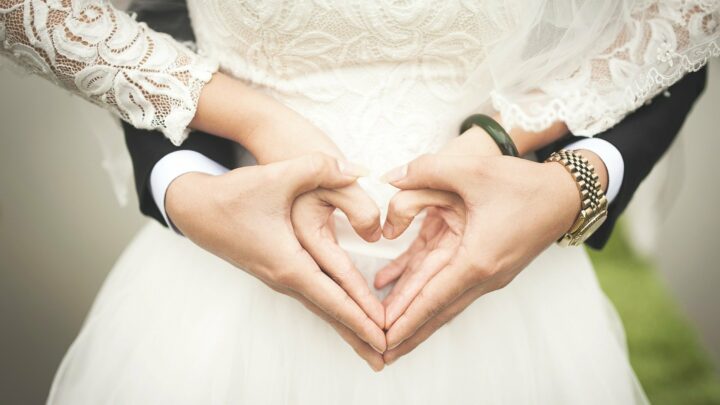 Zu sehen ist ein Brautpaar ohne Gesicht, die beide ein Herz mit ihren Händen formen.
